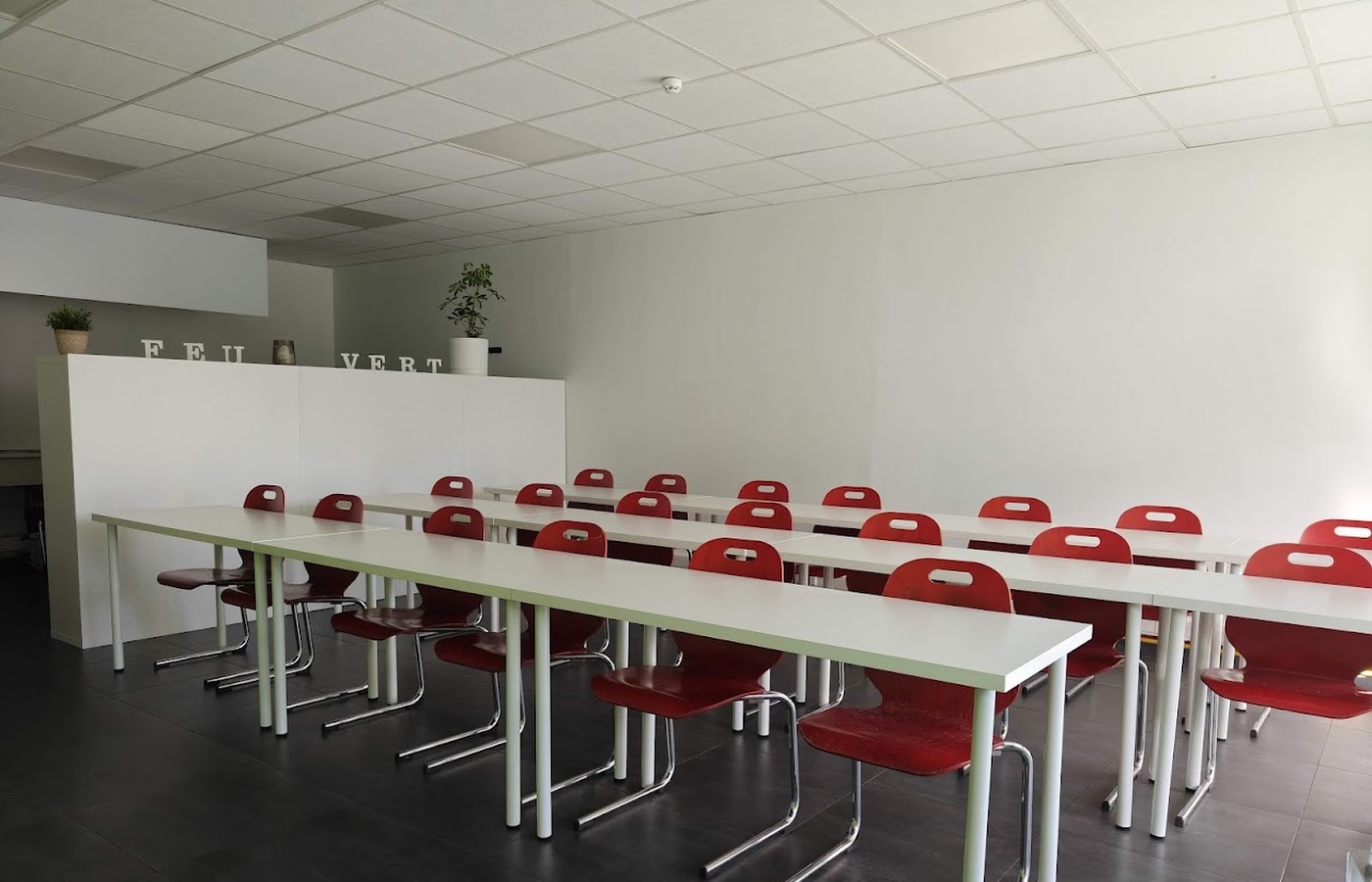 Salle de cours théoriques de l’auto-école Feu Vert Mons, rangées de tables blanches et de chaises rouges face au mur d’enseignement